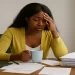 African American woman overwhelmed at a cluttered desk with papers and a coffee mug, illustrating how modern systems create exhaustion by design.