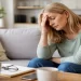 Woman sitting quietly on a couch at home, eyes closed and hand on her head, showing subtle signs of burnout and exhaustion