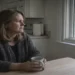 Middle-aged woman sitting alone at a kitchen table in early morning light looking down at her phone, reflecting the emotional weight of social isolation