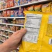 Hand holding a bag of ultra-processed snack food while reading the ingredient label in a grocery store aisle filled with packaged foods