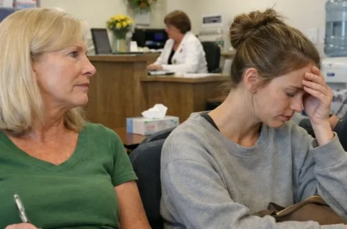 Two women sitting in a medical clinic waiting room, one holding paperwork and the other pressing her forehead in fatigue