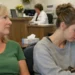 Two women sitting in a medical clinic waiting room, one holding paperwork and the other pressing her forehead in fatigue