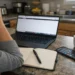 Woman sitting at kitchen counter looking overwhelmed with laptop, phone notifications, and blank notebook