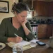 Woman writing in a notebook at a kitchen table with food, water, and a phone while tracking her health signals and daily patterns.