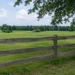 Weathered wooden fence beside a quiet green farmland field with a distant barn and AVFC logo, representing soil history, land stewardship, and rural legacy.