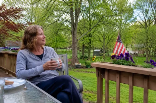 Woman sitting quietly on a backyard porch with coffee near an American flag, reflecting on health, freedom, and appreciation.