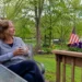 Woman sitting quietly on a backyard porch with coffee near an American flag, reflecting on health, freedom, and appreciation.