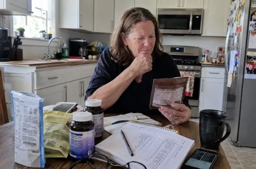 A woman sits at a kitchen table looking confused while comparing food labels, supplement bottles, notes, and health information, showing how conflicting health advice can feel overwhelming.