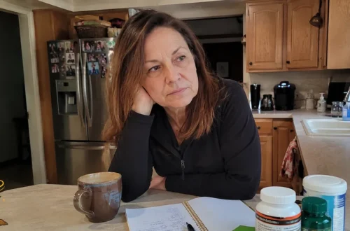Woman leaning at a kitchen counter with a notebook, coffee mug, fruit, and supplement bottles, looking thoughtful as she considers her choices.