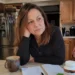 Woman leaning at a kitchen counter with a notebook, coffee mug, fruit, and supplement bottles, looking thoughtful as she considers her choices.