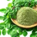 Moringa oleifera powder in a wooden bowl surrounded by fresh moringa leaves