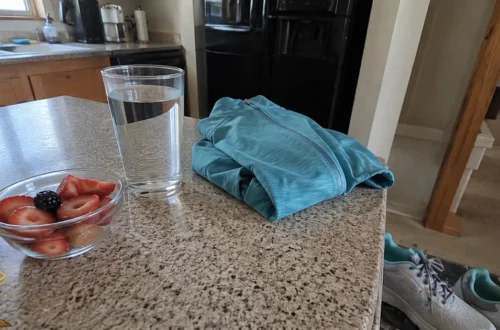 A simple kitchen counter with water, fresh fruit, a walking jacket, and sneakers representing steady healthy habits.
