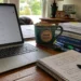 A realistic home office desk with a laptop showing health research results, handwritten notes, books, pens, highlighters, a mug, and the AVFC logo in the bottom corner.