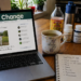 Kitchen table workspace with a laptop showing the A Voice For Change homepage, health supplements, tea, and a notebook with questions about root causes.