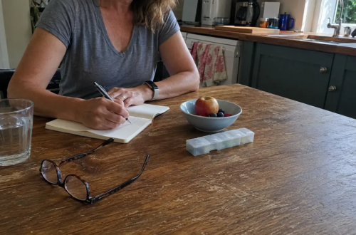 Woman writing health questions at her kitchen table beside water, fruit, glasses, and a pill organizer.