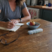 Woman writing health questions at her kitchen table beside water, fruit, glasses, and a pill organizer.