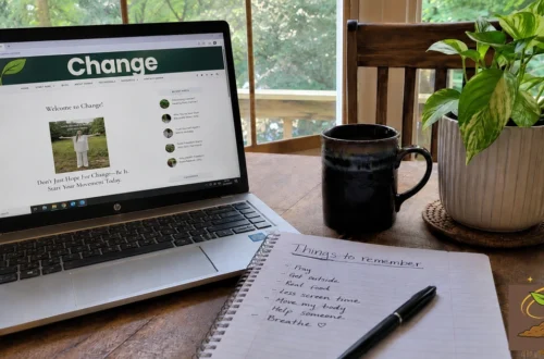 Laptop showing the A Voice For Change website beside a notebook, coffee mug, and houseplant in a natural home workspace