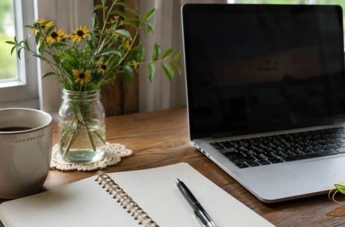 A quiet home desk with a laptop, notebook, pen, coffee mug, and flowers, representing a person taking responsibility for their own health questions and records.
