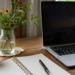 A quiet home desk with a laptop, notebook, pen, coffee mug, and flowers, representing a person taking responsibility for their own health questions and records.
