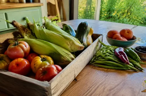 Fresh seasonal tomatoes, sweet corn, zucchini, peppers, peaches, and green beans arranged on a wooden kitchen table.